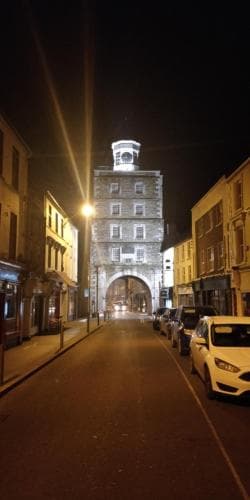A serene night scene captures a historic clock tower at the end of a quaint street lined with parked cars and illuminated by warm streetlights, ideal for travelers exploring charming European towns.
