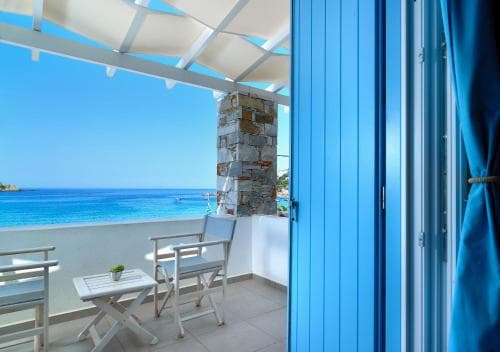 A serene coastal balcony with white furniture and a blue door frames a stunning view of the turquoise sea, ideal for travelers seeking relaxation in a picturesque seaside retreat.