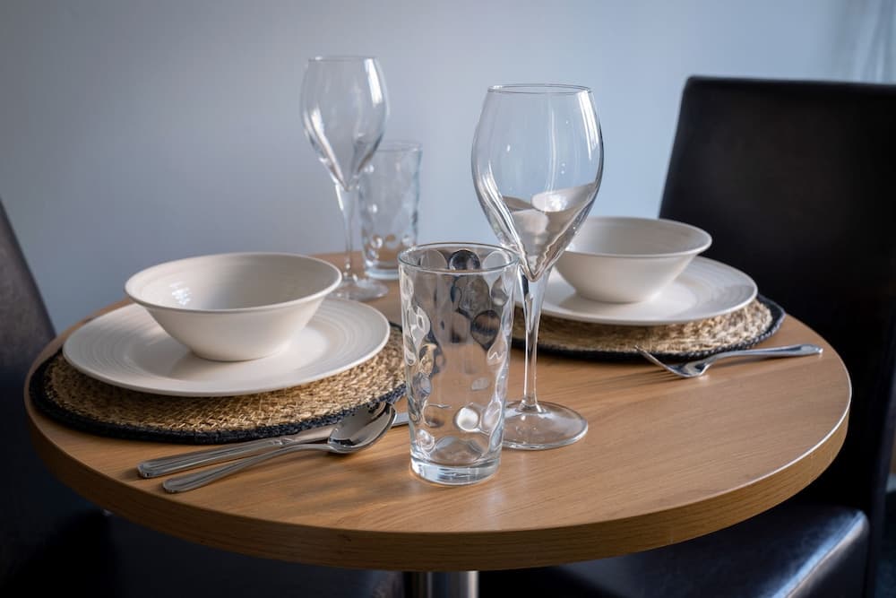 A cozy dining setup on a wooden table features elegant white bowls, plates, wine glasses, and textured placemats, ideal for a romantic dinner or intimate gathering in a modern restaurant setting.