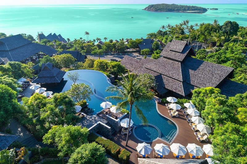 An aerial view of a luxurious resort nestled among lush greenery with a stunning infinity pool overlooking the turquoise sea, perfect for travelers seeking a serene tropical getaway.