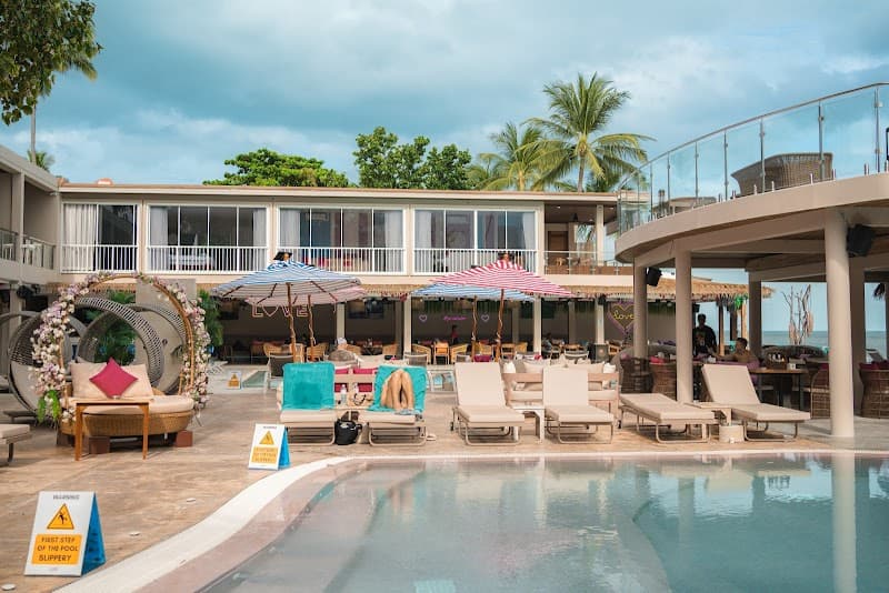 A vibrant beach club scene featuring lounge chairs, colorful umbrellas, and a poolside area surrounded by lush greenery and palm trees, ideal for travelers seeking relaxation and tropical ambiance in Koh Samui, Thailand.