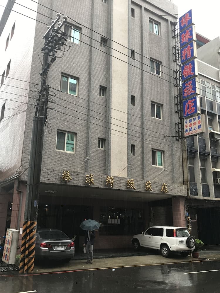 A rainy urban street scene features a multi-story building with Chinese signage, parked cars, and a person holding an umbrella, ideal for travelers seeking local dining or accommodation options in a bustling city environment.