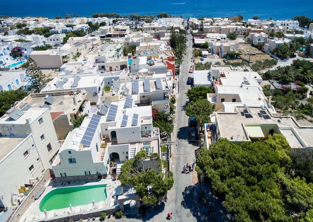 An aerial view of a sunlit coastal town with white buildings, solar panels, lush greenery, and a vibrant pool, ideal for travelers seeking a serene Mediterranean retreat.