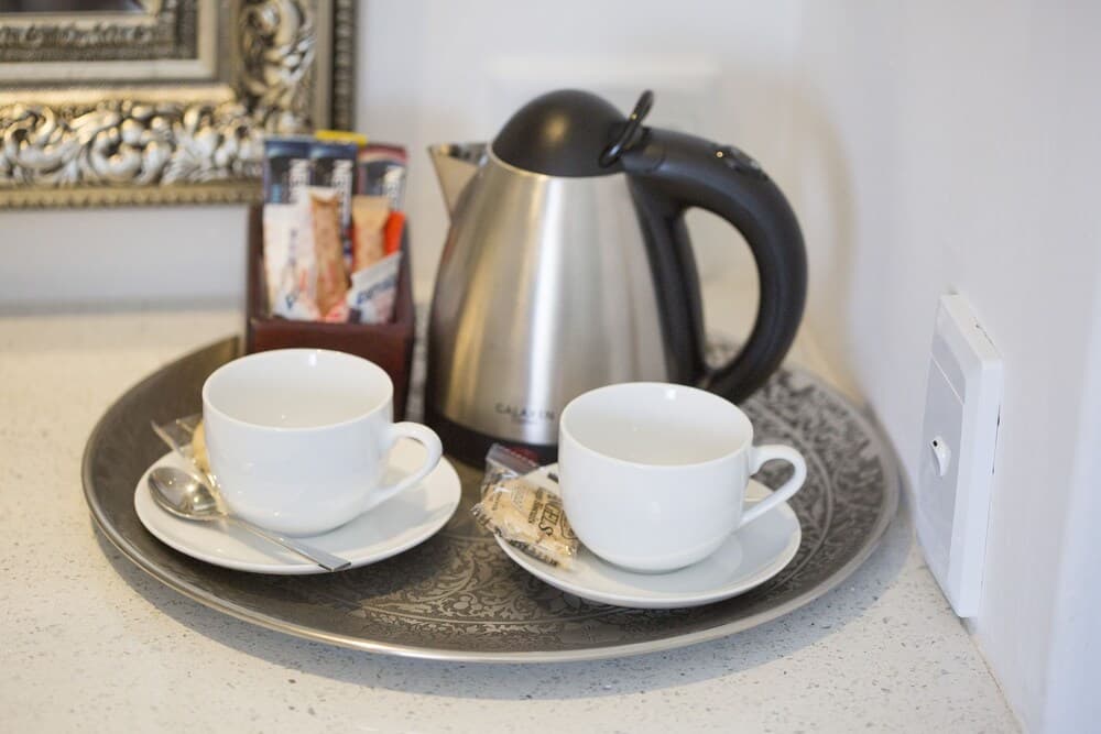 A cozy hotel room corner features a tray with two white cups, saucers, a stainless steel kettle, sugar packets, and a decorative mirror in the background, ideal for travelers seeking a comfortable tea or coffee break.