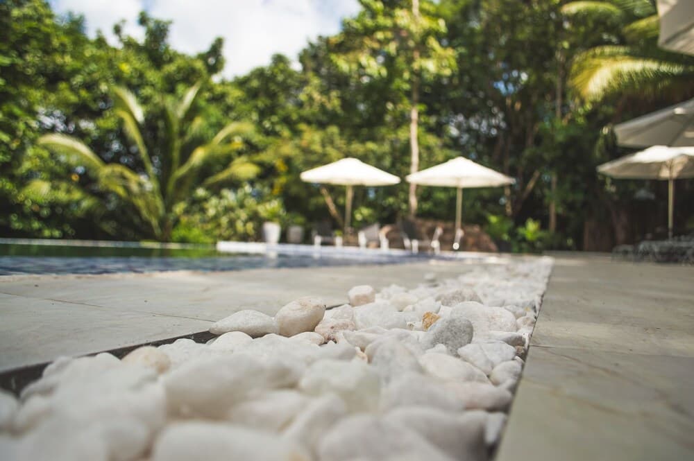 A serene tropical poolside scene with white pebbles, lush greenery, and white umbrellas creates an inviting atmosphere ideal for relaxation and vacation photography.