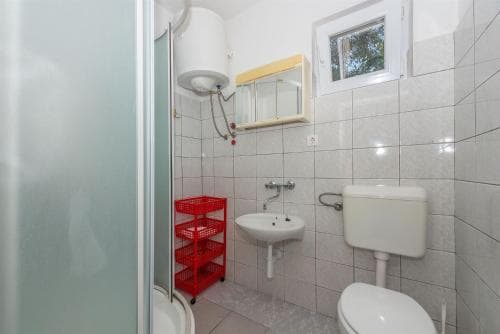 A compact bathroom with tiled walls, a sink, toilet, shower area, and red storage cart offers a practical space for travelers seeking basic amenities in a functional setting.