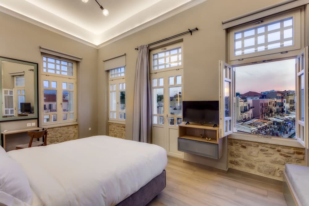 A cozy hotel room with a neatly made bed, wooden floors, and large windows offering a picturesque view of a bustling cityscape at sunset, ideal for travelers seeking comfort and scenic vistas.
