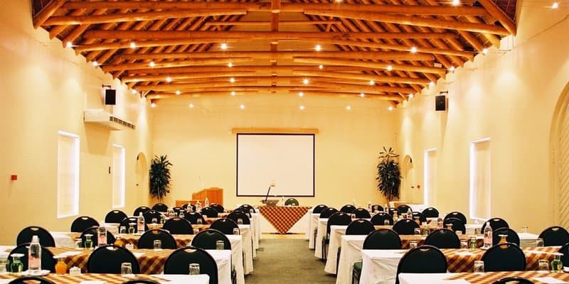 A spacious conference hall with wooden beams, warm lighting, and neatly arranged tables and chairs creates an inviting atmosphere ideal for professional gatherings and events.