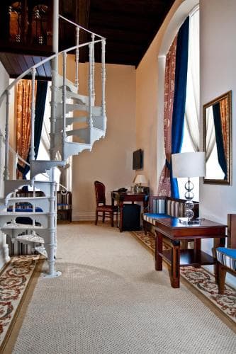 A cozy hotel room features a spiral staircase leading to an upper level, warm lighting, patterned rugs, and elegant furnishings, creating an inviting atmosphere perfect for travelers seeking comfort and charm.