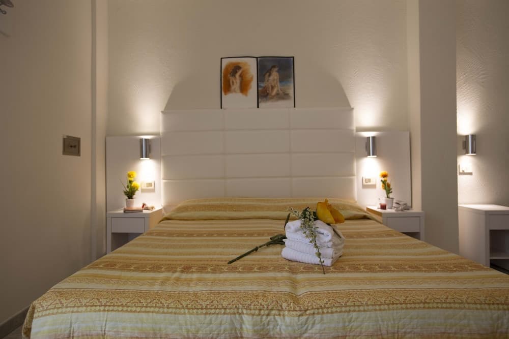 A cozy hotel bedroom features a neatly made bed with yellow bedding, white towels artfully arranged on top, soft wall lighting, and decorative paintings above the headboard, ideal for travelers seeking comfort and relaxation.