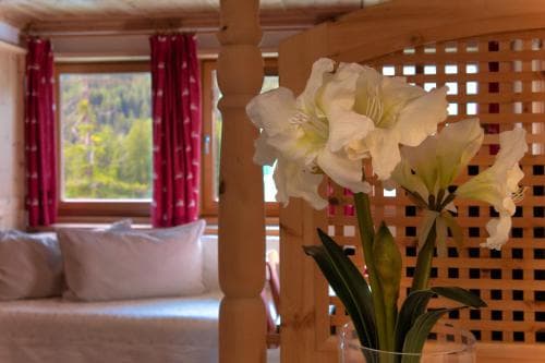 A cozy cabin interior features a vase of white flowers on a wooden table, framed by red curtains and a window view of lush greenery, ideal for a serene getaway retreat.
