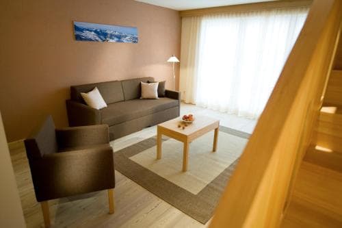 A cozy living room with a sofa, armchair, wooden coffee table, and mountain artwork, bathed in soft light from sheer curtains, offers a serene retreat ideal for travelers seeking relaxation in a modern lodge setting.