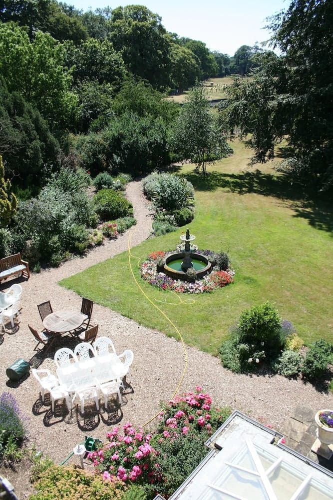 A serene garden with a central fountain surrounded by lush greenery, vibrant flower beds, and outdoor seating areas, ideal for relaxation and leisurely strolls.