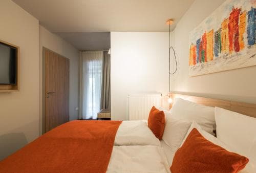 A cozy hotel room features a neatly made bed with orange accents, modern decor, and vibrant cityscape artwork, ideal for travelers seeking comfort and style during their stay.