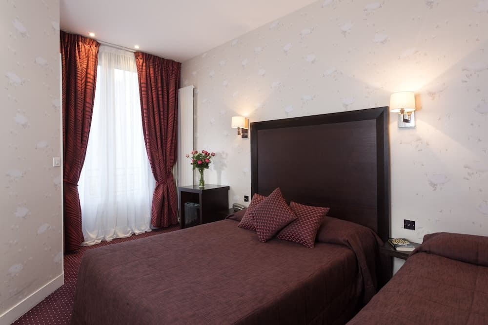 A cozy hotel room features a neatly made bed with maroon bedding, floral wallpaper, red curtains, and bedside lamps, ideal for travelers seeking comfort and relaxation.