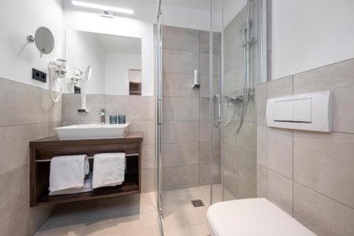 A modern hotel bathroom features sleek tiled walls, a glass-enclosed shower, a wooden vanity with neatly folded towels, and a minimalist design, ideal for travelers seeking comfort and cleanliness.