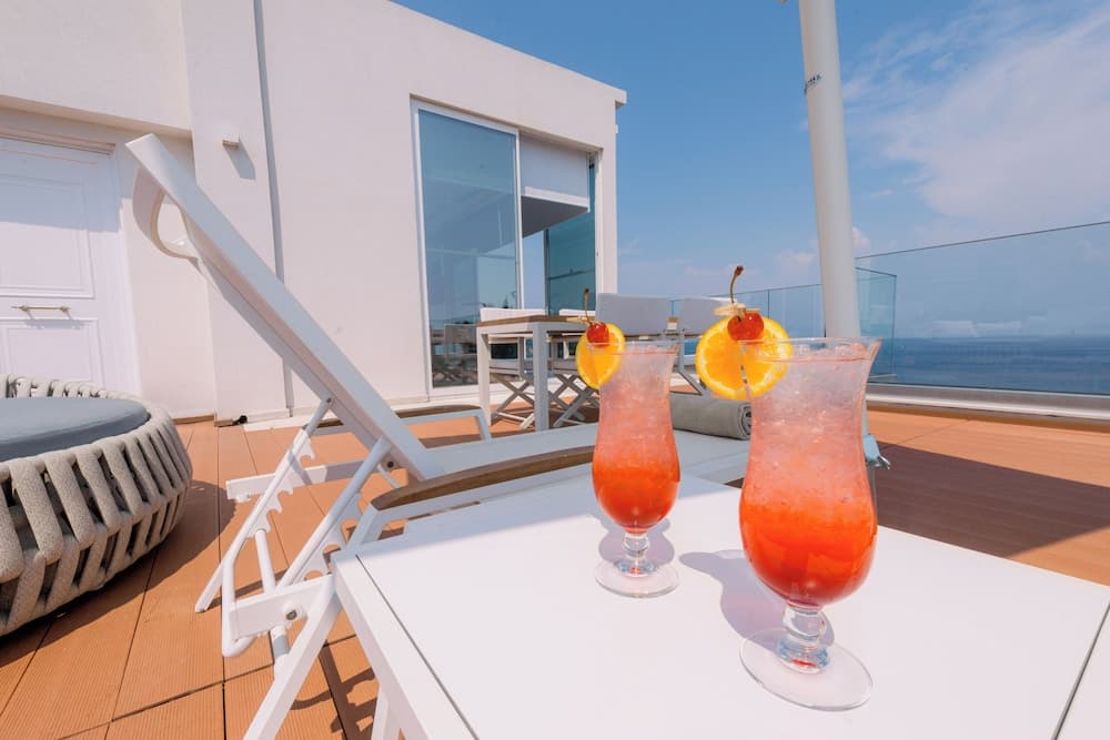 A serene rooftop terrace features vibrant cocktails on a white table, modern lounge chairs, and a stunning ocean view, perfect for a relaxing getaway or vacation rental promotion.