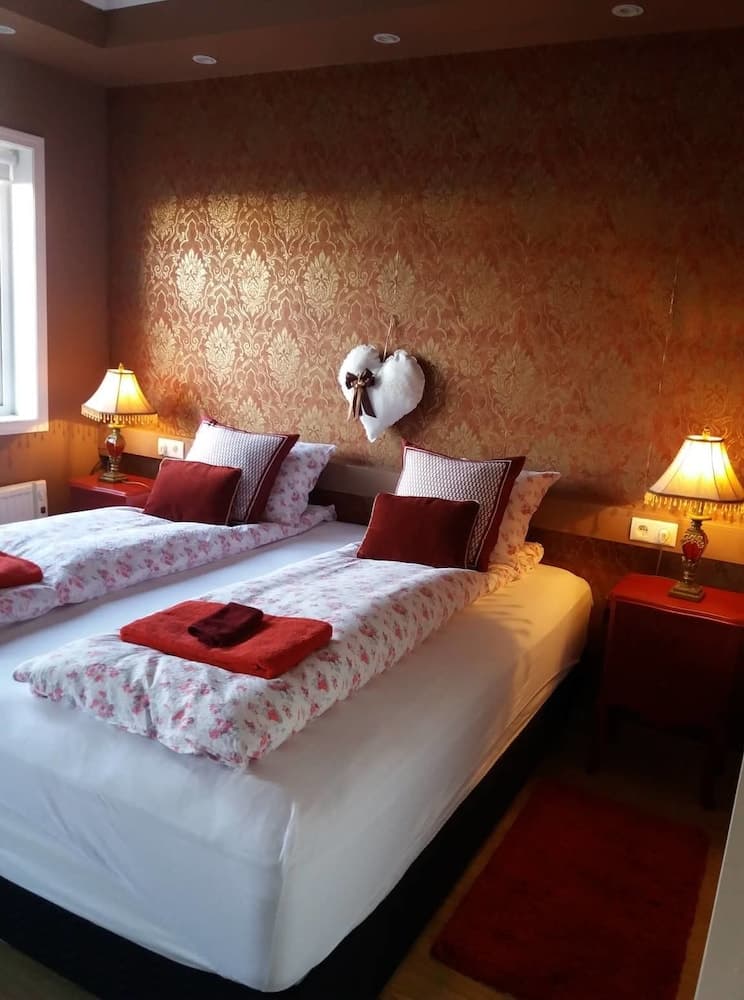 A cozy hotel room features twin beds adorned with floral bedding and red accents, warm lighting from bedside lamps, and a decorative heart on the patterned wall, ideal for travelers seeking comfort and charm.