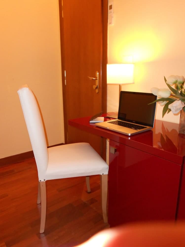 A cozy hotel room workspace features a sleek red desk with a laptop, white chair, soft lighting from a lamp, and fresh flowers, ideal for travelers seeking comfort and productivity.