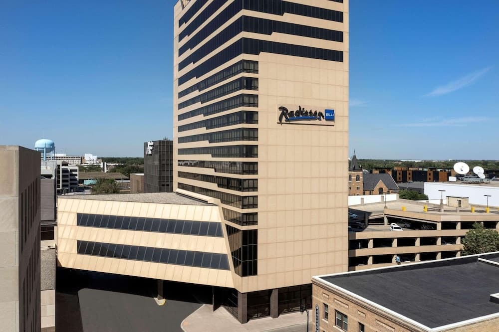 A modern Radisson Blu hotel stands tall against a clear blue sky in an urban setting, surrounded by parking structures and other buildings, ideal for business travelers seeking convenience and comfort.