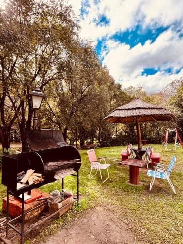 A serene outdoor picnic area features a barbecue grill, colorful chairs under a thatched umbrella, surrounded by lush greenery and a clear blue sky, perfect for a relaxing family getaway or camping adventure.
