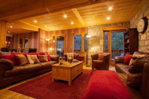 A cozy cabin living room with warm wooden interiors, plush red sofas, and stone walls exudes comfort, perfect for travelers seeking relaxation in a rustic retreat.