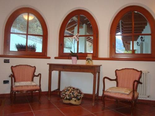 A cozy indoor lounge with arched wooden windows overlooking greenery, featuring two cushioned chairs, a central table adorned with flowers, and warm terracotta flooring, ideal for a relaxing retreat or vacation rental setting.