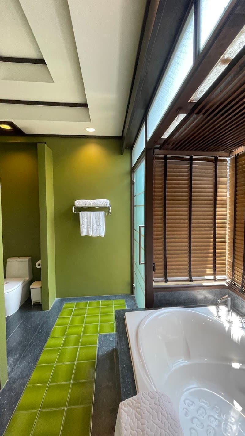 A serene bathroom at Nora Buri Resort & Spa on Koh Samui features a modern bathtub, green-tiled floor, wooden blinds, and plush towels, ideal for travelers seeking relaxation and luxury.