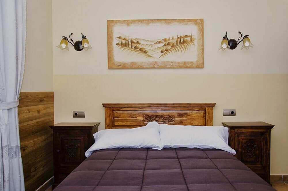 A cozy bedroom with a wooden bed, matching nightstands, wall-mounted lamps, and a serene landscape painting creates a tranquil atmosphere ideal for travelers seeking relaxation in a rustic retreat.