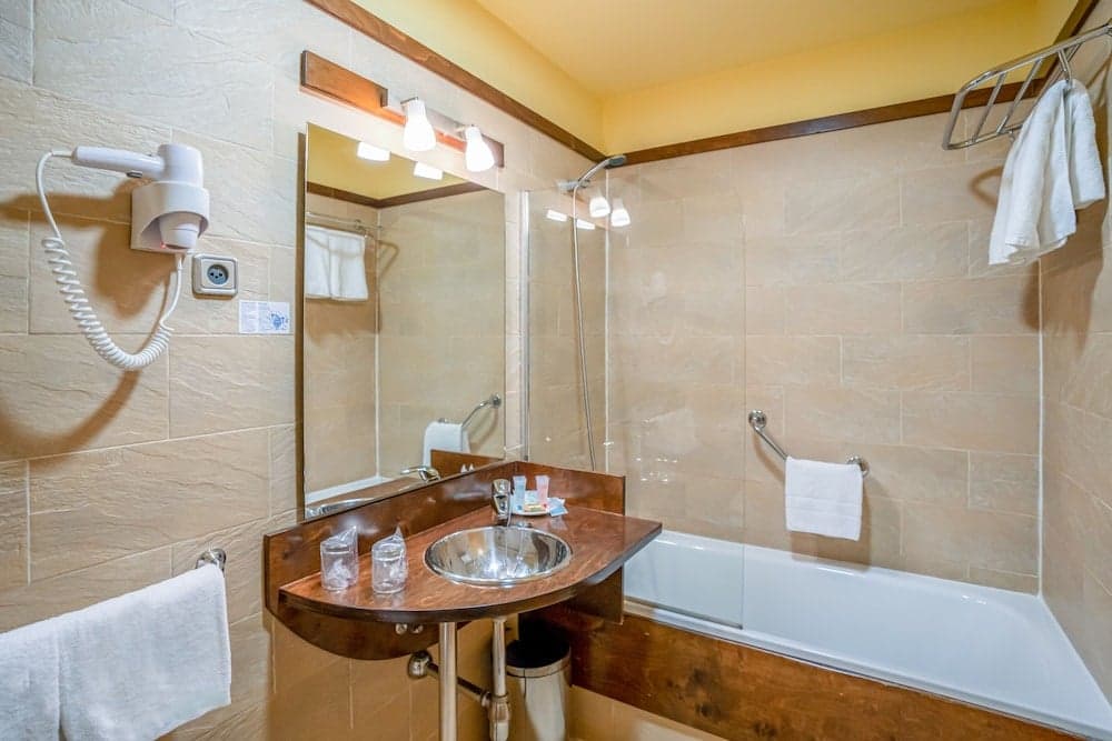 A cozy hotel bathroom features beige tiled walls, a wooden vanity with a sink, a bathtub with a shower, neatly hung white towels, and a wall-mounted hairdryer, ideal for travelers seeking comfort and convenience.