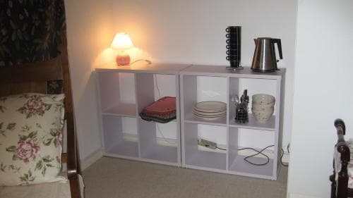A cozy bedroom corner features white shelving units holding decorative items like plates, cups, and a kettle, illuminated by a soft lamp, ideal for travelers seeking a comfortable and personalized space.