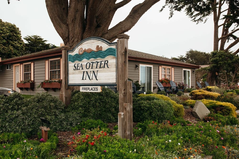 A charming coastal inn surrounded by lush greenery and a welcoming sign featuring a sea otter motif invites travelers seeking a cozy retreat with fireplaces.