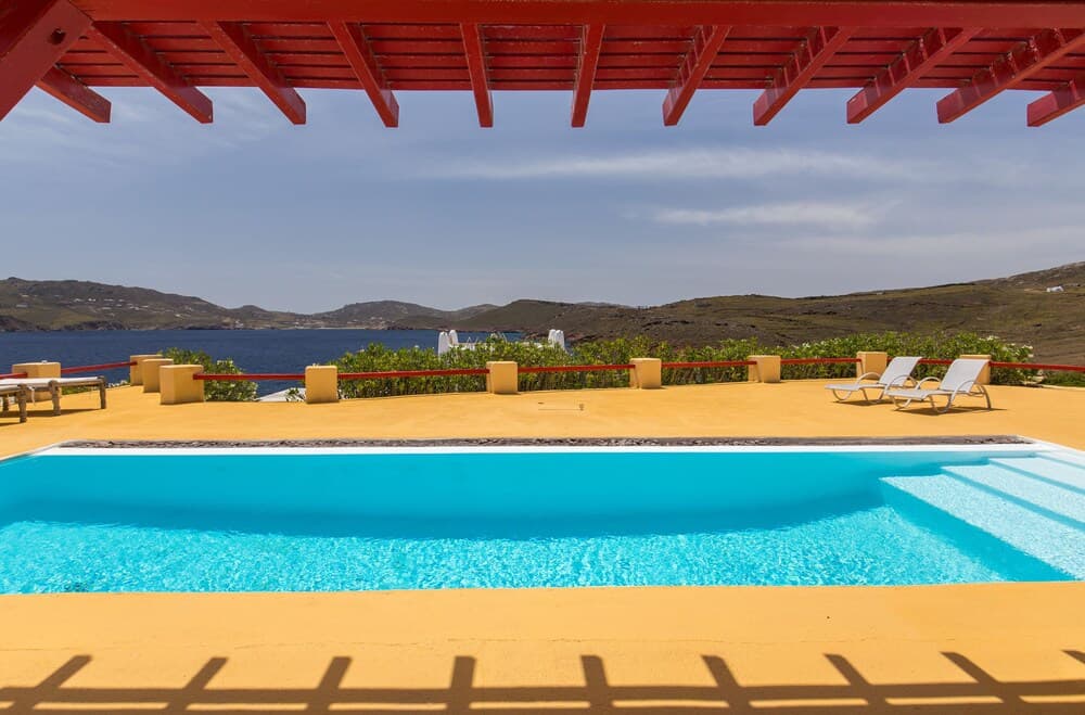 A serene coastal villa features a vibrant blue pool under a red pergola, surrounded by lounge chairs and overlooking the tranquil sea and distant hills, ideal for luxury retreats.