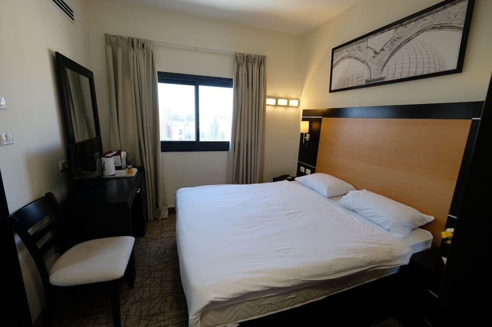 A cozy hotel room features a neatly made bed with white linens, a wooden headboard, a vanity with a mirror, and a window with curtains, creating a serene atmosphere ideal for travelers seeking comfort and relaxation.