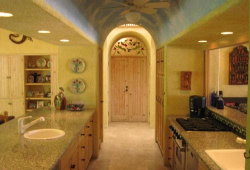 A warmly lit kitchen with vibrant yellow walls, decorative plates, and an inviting arched doorway leads to a cozy interior, perfect for travelers seeking a homely culinary retreat.