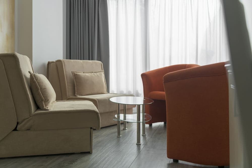 A cozy hotel room features a beige sofa, orange armchair, glass coffee table, and soft natural light through sheer curtains, creating a serene atmosphere ideal for relaxation during travel.