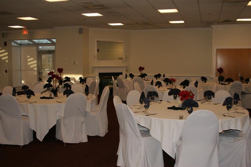 A well-lit banquet hall set up for an elegant event features round tables draped in white linens, adorned with red floral centerpieces and black napkins, ideal for wedding receptions or corporate gatherings.