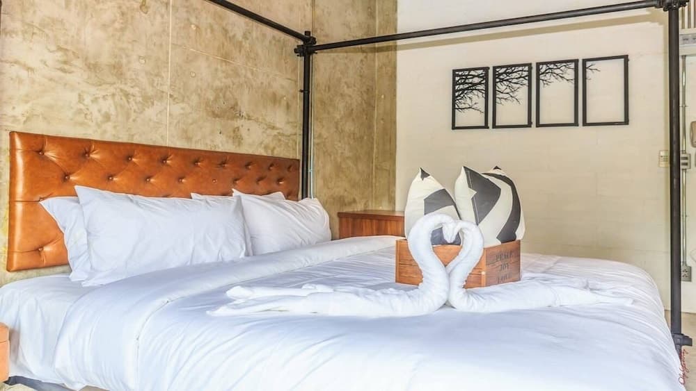 A cozy hotel room features a plush bed with white linens, a leather headboard, decorative pillows, and towel swans, set against textured walls and framed tree art, ideal for travelers seeking comfort and charm.