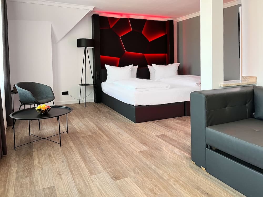 A modern hotel room features a cozy ambiance with a striking red-lit geometric headboard, sleek furniture including a gray sofa and chair, and a fruit bowl on a table, ideal for travelers seeking comfort and style.