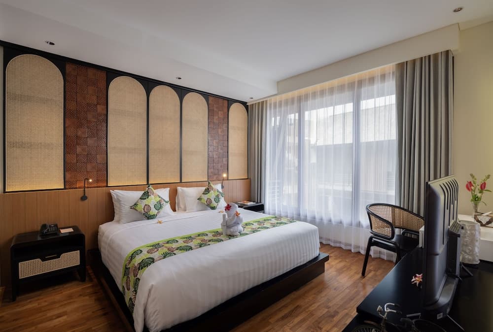 A cozy hotel room with a neatly made bed adorned with tropical-themed pillows and a decorative runner, featuring a stylish woven partition wall, large window with sheer curtains, and modern furnishings, ideal for travelers seeking comfort and relaxation.