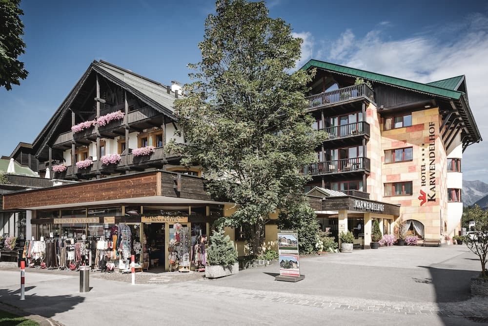 A charming alpine hotel with balconies adorned with pink flowers stands beside a quaint shop under a clear blue sky, ideal for travelers seeking a cozy mountain retreat.