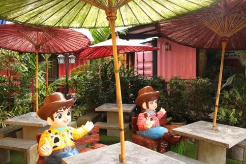 A vibrant outdoor café adorned with colorful parasols and playful cowboy-themed statues invites travelers to enjoy a whimsical and relaxing dining experience amidst lush greenery.