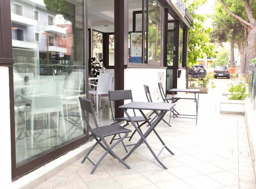 A serene outdoor café patio features modern folding chairs and tables under large glass windows, surrounded by lush greenery, ideal for travelers seeking a quiet spot to relax and enjoy refreshments.