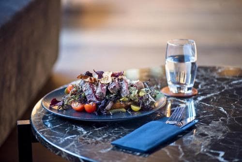 A beautifully plated salad with vibrant vegetables sits on a sleek marble table beside a glass of water, evoking a sophisticated dining experience ideal for travelers seeking upscale culinary adventures.