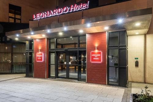 A warmly lit entrance to the Leonardo Hotel at night, featuring modern architecture with glass doors and red brick walls, creates an inviting atmosphere ideal for travelers seeking comfortable accommodations.
