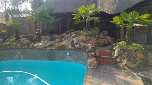 A serene tropical resort poolside scene features a vibrant blue pool surrounded by lush palm trees, natural rock formations, and a thatched-roof structure, ideal for relaxing vacations and leisurely getaways.