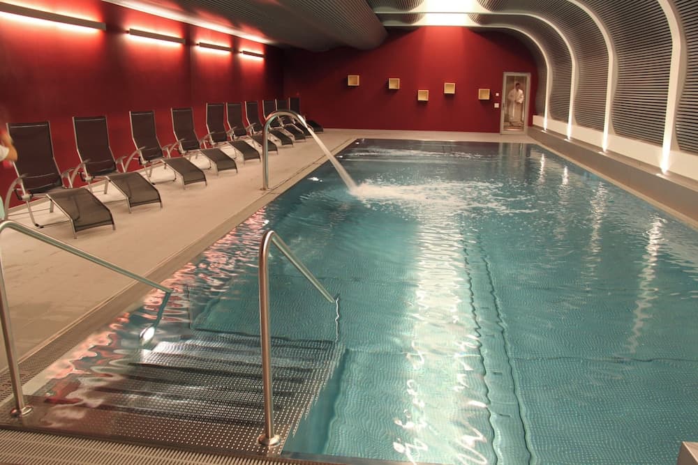 A serene indoor pool area with red walls, lounge chairs, and a waterfall feature offers a relaxing retreat for spa-goers seeking tranquility and rejuvenation.