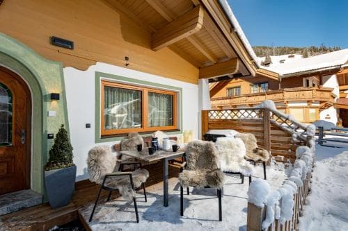 A cozy outdoor seating area with fur-covered chairs and a wooden table is nestled in a snowy alpine setting, perfect for travelers seeking a serene winter retreat.