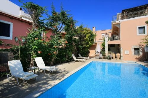 A serene Mediterranean courtyard features a sparkling pool surrounded by lounge chairs, lush greenery, and colorful buildings, ideal for a relaxing vacation retreat.