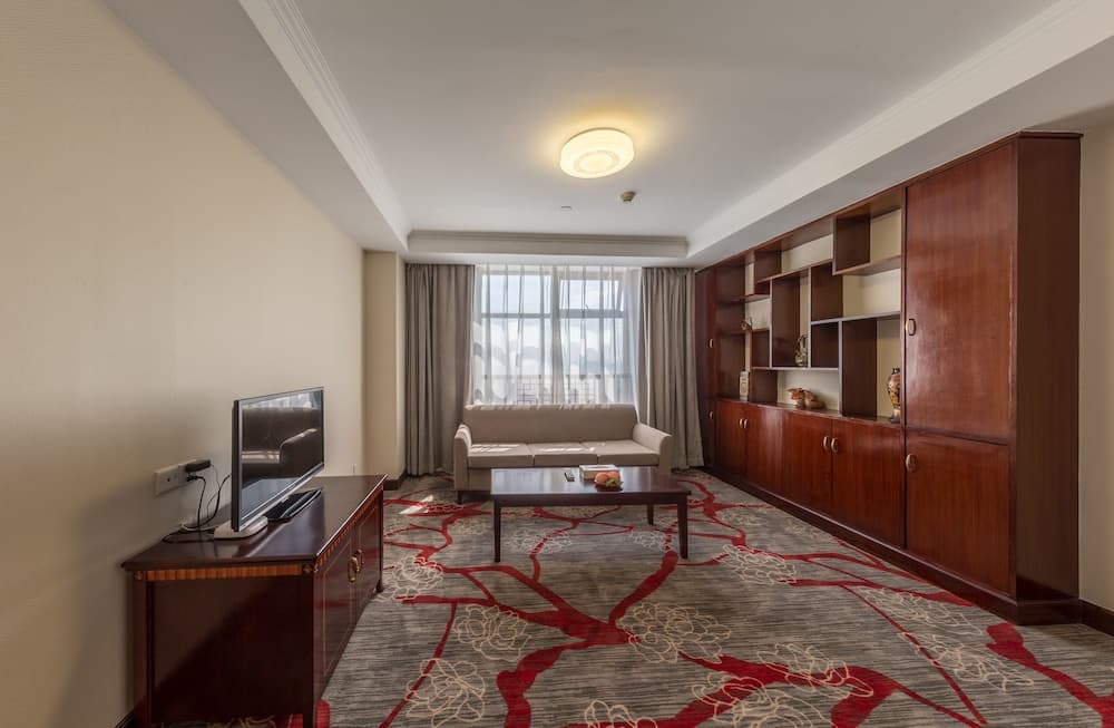 A cozy hotel suite features a plush sofa by a large window, a wooden entertainment unit with a TV, and a stylish carpet, ideal for travelers seeking comfort and relaxation.